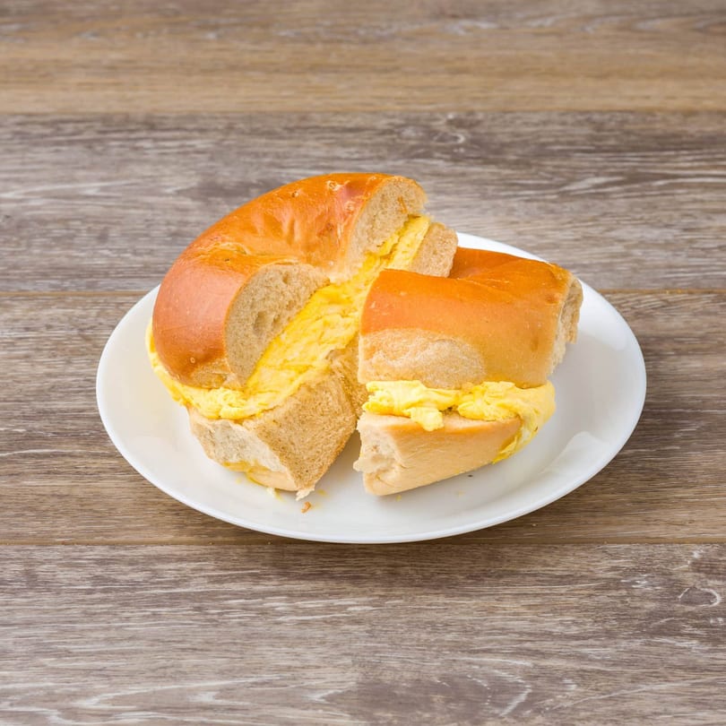 A sliced bagel with scrambled eggs on a white plate, on a wooden table.