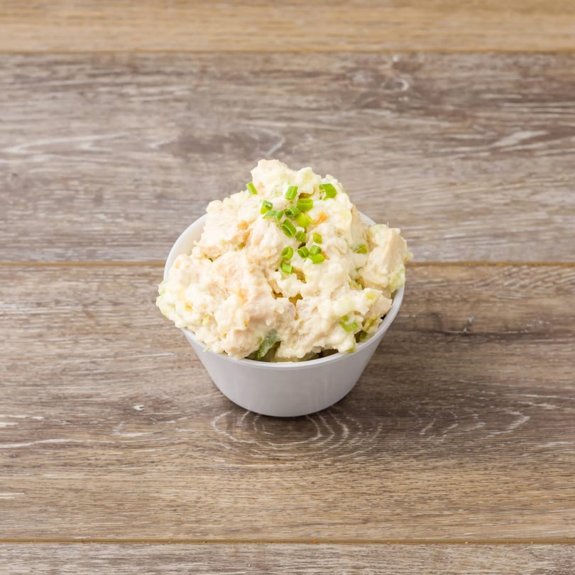 A white bowl of creamy potato salad, topped with fresh green chives, on a light wooden surface.